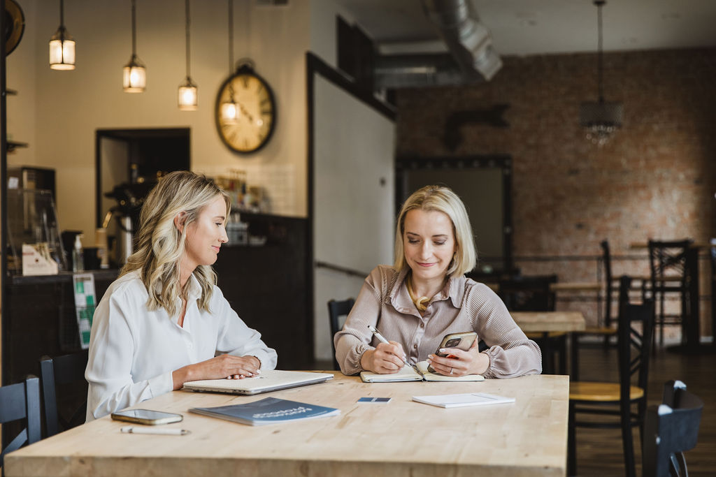 Two people collaborating in a meeting while one person is taking notes in a journal while using a phone.